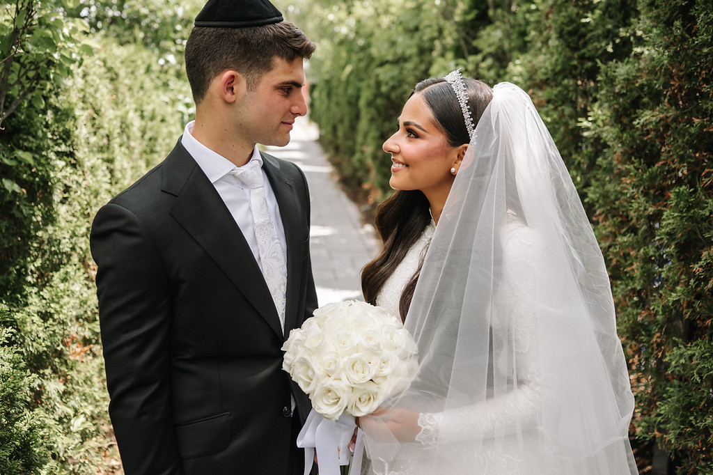 Frum bride and groom in black and white attire outdoors, standing close and looking at each other with the bride’s veil and bouquet in view.