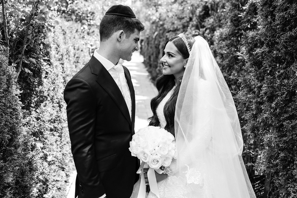 Black-and-white wedding portrait of the groom and bride facing each other on a garden path, the bride smiling beneath her veil and holding a bouquet.