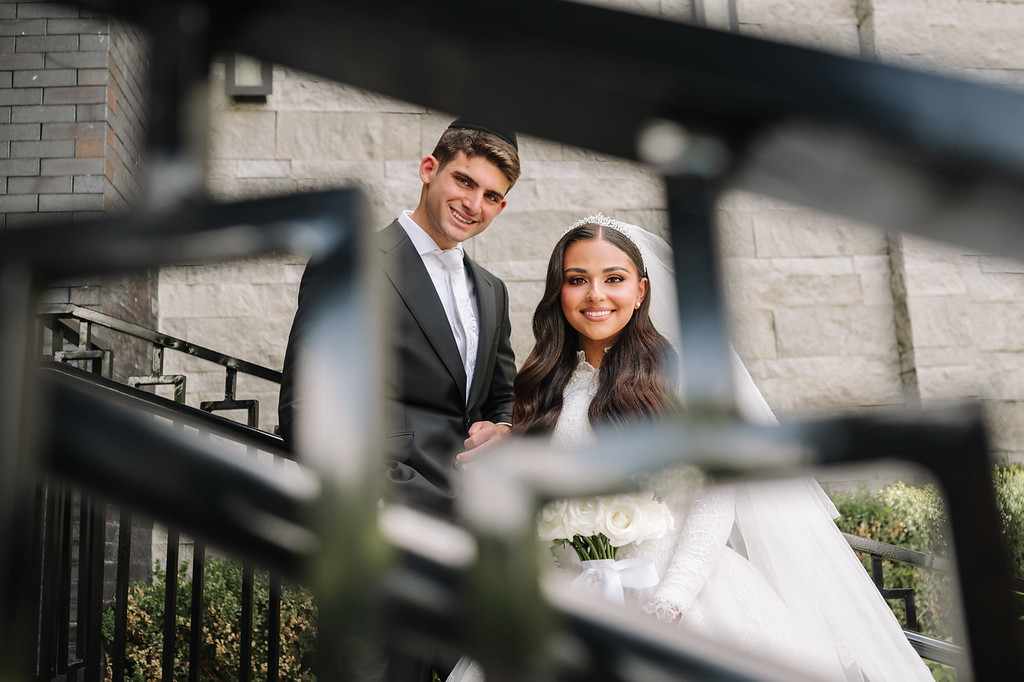 Smiling bride in a white gown and veil with groom standing behind her outdoors in front of stone buildings and a softly blurred foreground.