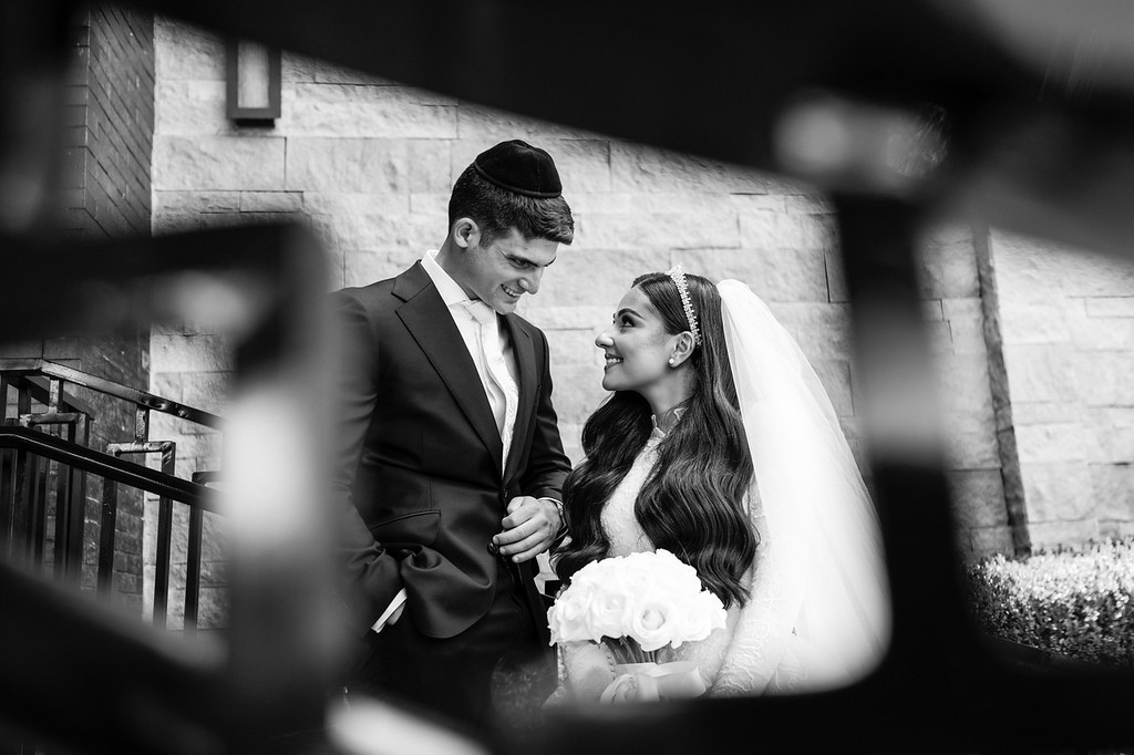 Black-and-white photo of the groom and kallah during the badeken, smiling at each other with clasped hands as the bride is covered with a veil.