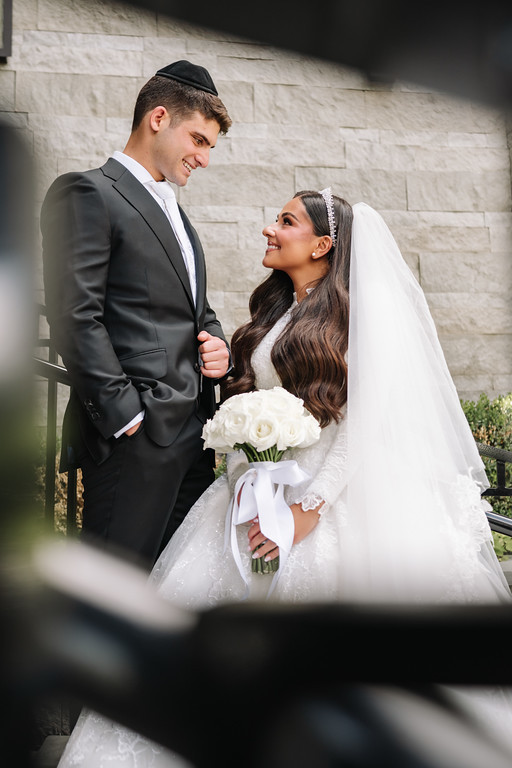 Bride and groom in formal wedding attire smiling at each other during a close embrace, with a white bouquet in the bride’s hands and stone wall background.