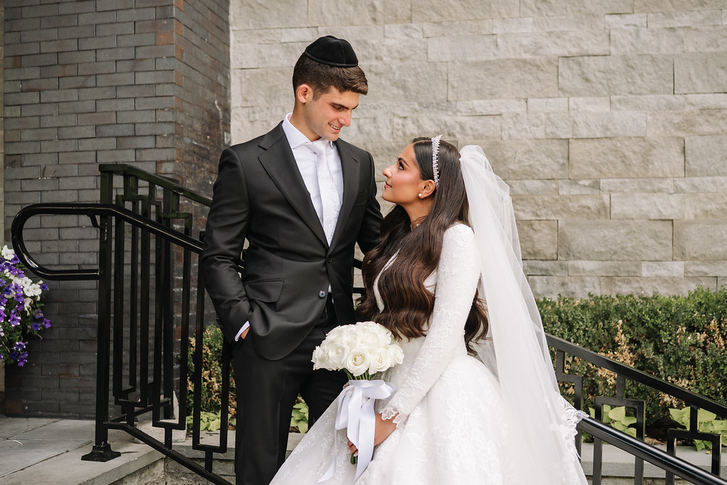 Bride and groom in formal wedding attire share an intimate moment outdoors against a stone wall, smiling as they look at each other.