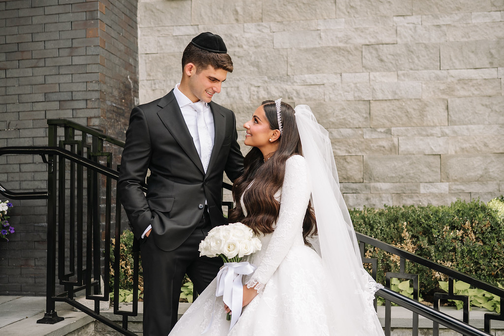 Frum wedding couple smiling at each other during portraits, the groom in a black kippah and suit and the bride in a white gown holding a bouquet by a stone wall and railing.