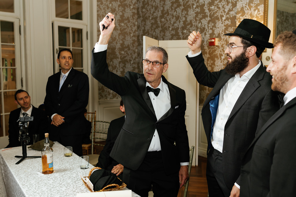 An older man in a black suit and hat raises both hands in celebration at a seated banquet table as other guests look on.