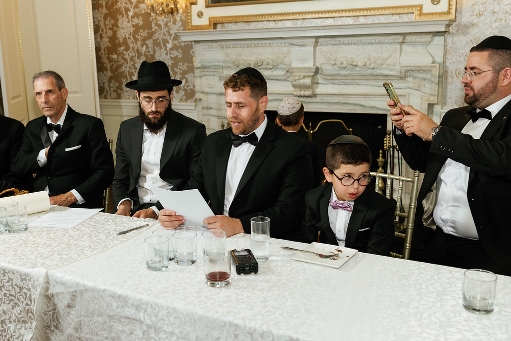 Men in black hats and bowties seated at a decorated venue table during a formal moment, with a young boy in glasses in the foreground and one attendee checking a phone.