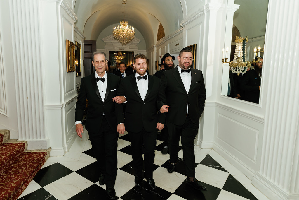 Four men in black tuxedos with bow ties pose in a grand chandelier-lit hallway on a black-and-white checkered floor.