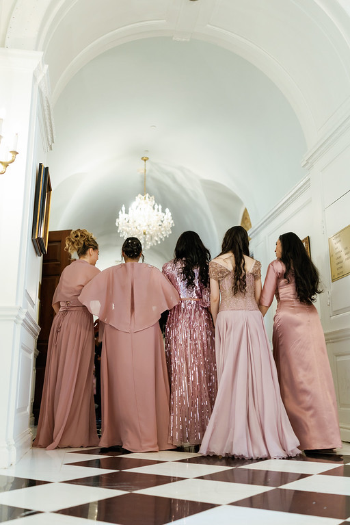 A group of bridesmaids and guests in blush matching dresses stand and walk together down a grand hallway beneath a chandelier, viewed from behind.