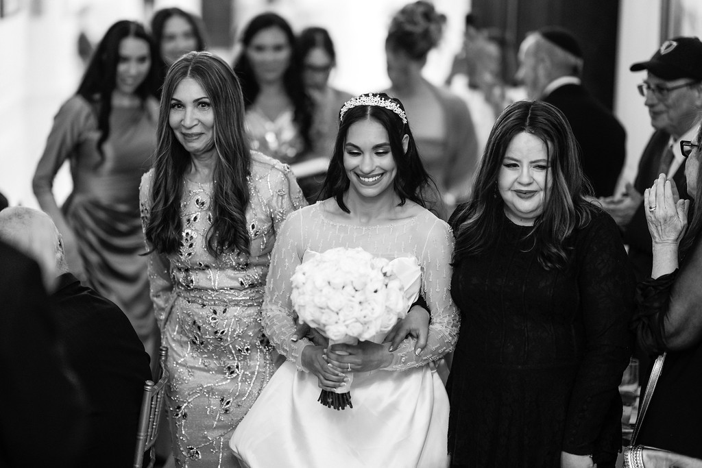 Black-and-white wedding photo of the bride walking forward holding a bouquet, with her mother beside her and guests watching in the background.