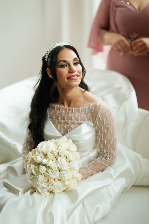 Frum bride in an ivory wedding dress seated on a white chair, holding a white rose bouquet, looking toward the camera with a soft smile.