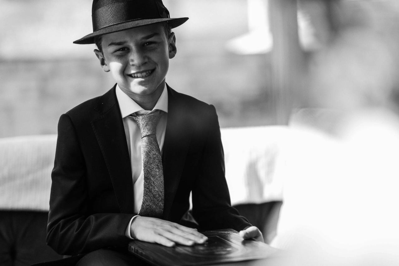 Black-and-white portrait of a smiling bar mitzvah boy wearing a dark fedora and suit, seated indoors with a softly blurred background.