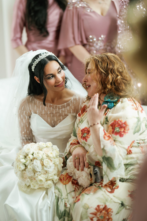 Bride in a white gown and jeweled headpiece smiles warmly during kallah/greeting moment with an older woman leaning in beside her, floral bouquet in the foreground, wedding guests blurred in the background.