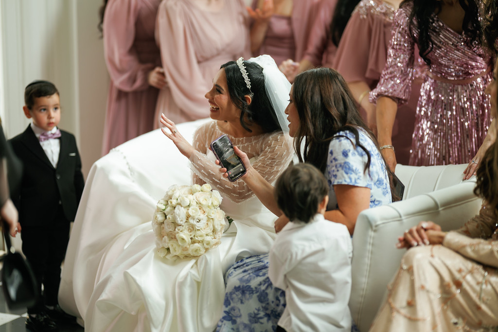 Bride in a white gown sits on a sofa with family and guests nearby; a young boy in blue looks toward her while guests chat and react.