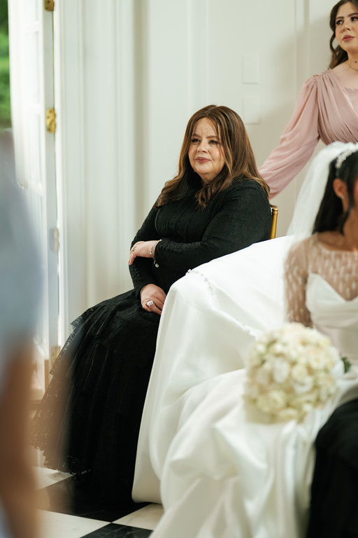A woman in a black outfit sits on a gold chair during a wedding moment, with a bride in the foreground and guests in the background.