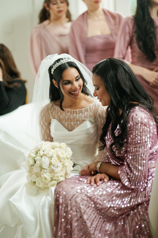 Frum bride in a white lace-trim veil laughs as a young woman in a pink sequined outfit leans in with her hands near the bride’s forearm, with bridesmaids seated in the background.