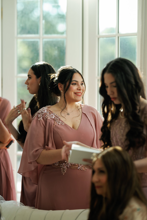 Bride in a blush pink gown with jewelry and a gentle smile stands by a group of bridesmaids in a sunlit room with large windows.