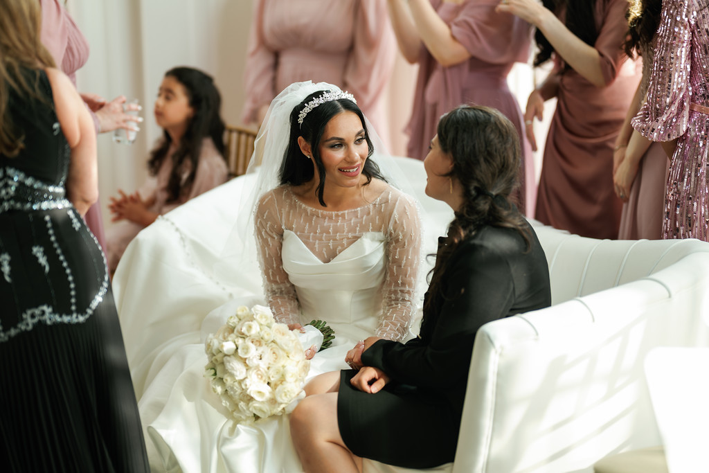 Frum bride wearing a lace veil and tiara-style headpiece sits in a white chair holding a bridal bouquet while a guest in black leans in beside her during the celebration.