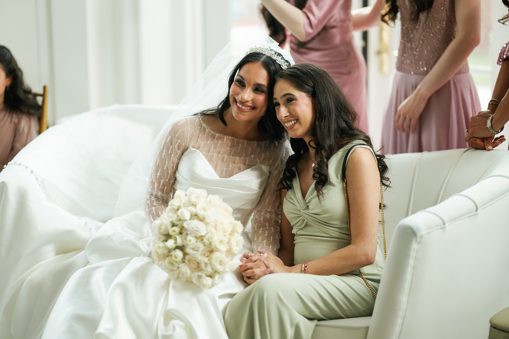 Smiling bride in a white dress sits on a cushioned sofa with a close companion in a light green dress; soft bridal details and white floral bouquet are visible during the wedding reception.