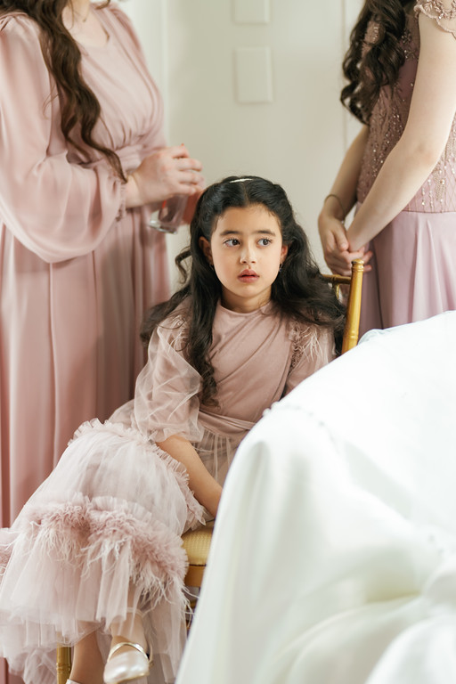 A young woman stands in the center during badeken, looking up solemnly as women in pale pink surround her indoors.