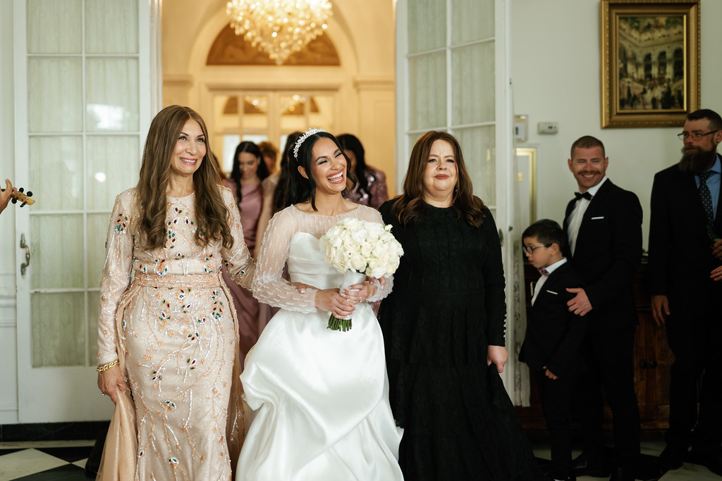 Bride in a white dress and veil smiles while holding a bouquet, walking beside family members inside an elegant hall with warm chandelier light.