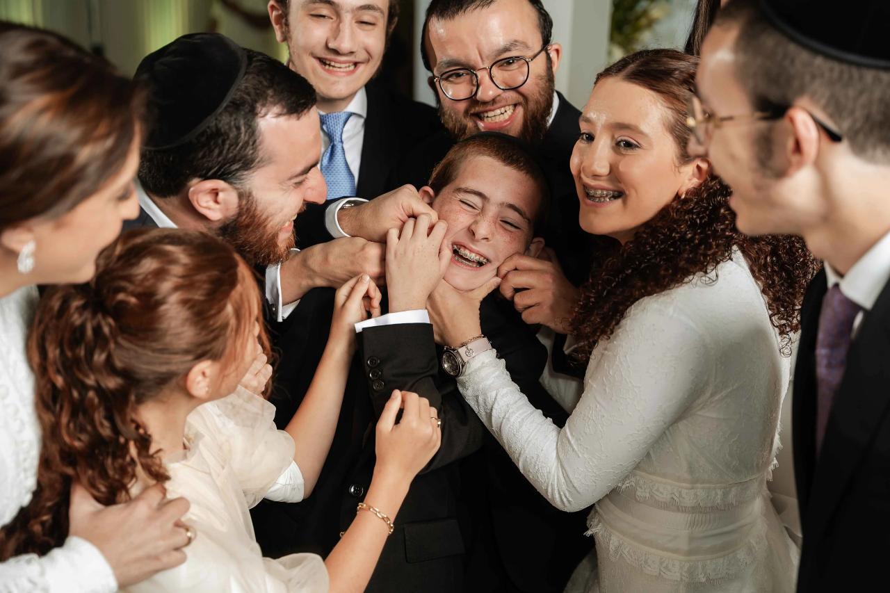 Smiling Jewish wedding guests surround a laughing child in the middle of a joyful celebration as everyone leans in and claps.