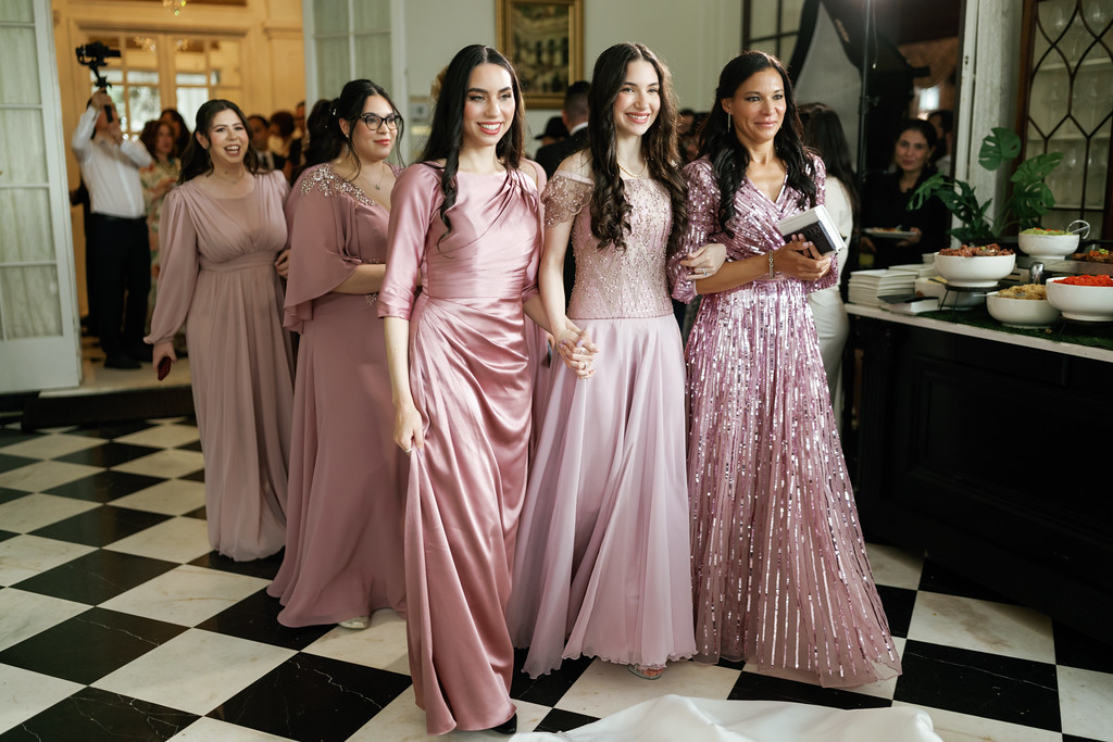 The bride in a pink satin gown walks hand-in-hand with bridesmaids in matching pink dresses in a decorated ballroom hallway.