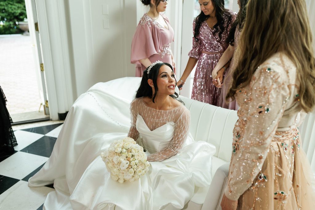 A bride in a white gown lounges on a white chair holding a bouquet while guests stand nearby in formal dresses.