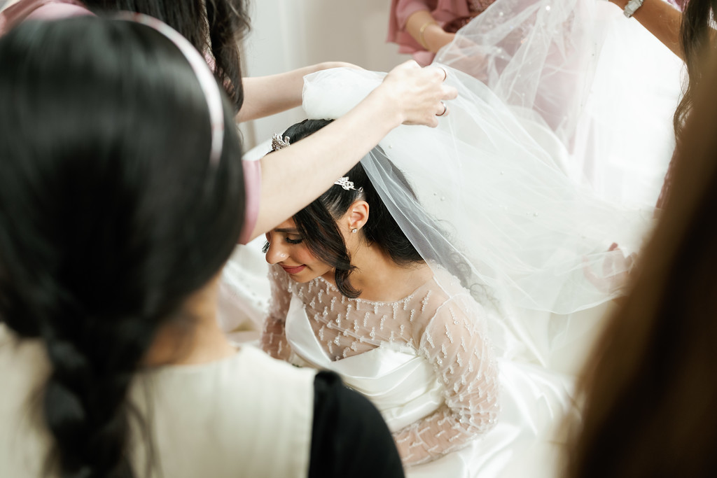 The bride looks down while family members adjust her bridal veil during badeken, surrounded by guests in a soft, bright setting.