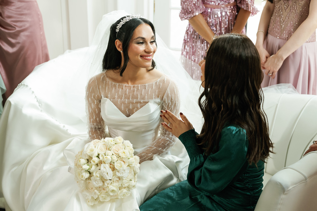 Bride smiling during the bedeken as a woman adjusts her veil, with the bride holding a white rose bouquet; guests in pastel dresses stand in the background.