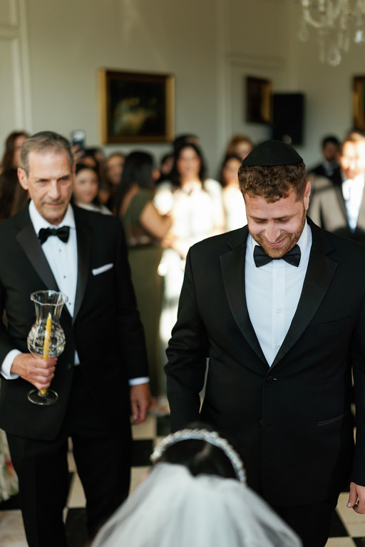 A groom in a black suit and kippah laughs with his head bowed, standing at a formal event while a second man in a bow tie holds a drink behind him.