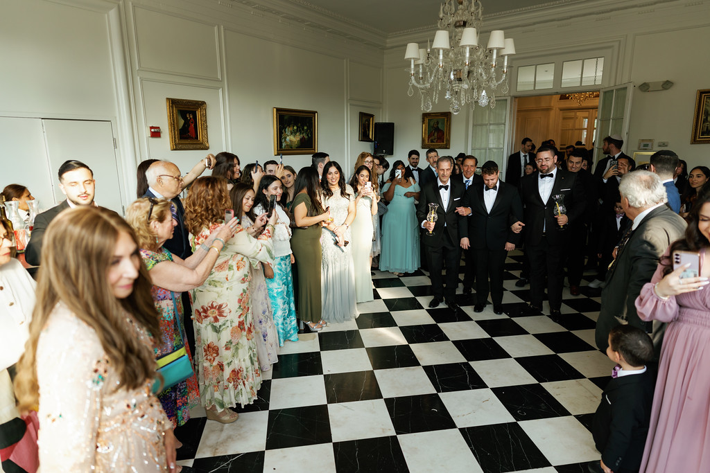 A wide ballroom scene where guests in formal attire dance and clap, with the groom and attendants in dark suits along the right side near a line of guests.