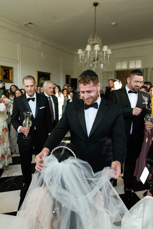 At a wedding in a reception hall, the groom in a black kippah and tuxedo lifts the bride’s veil while guests in formalwear watch from behind.