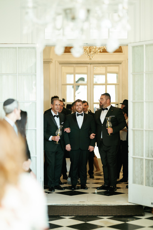 Three men in black tuxedos with bow ties and two chalices/goblets move through a ballroom doorway while smiling, as other guests look on behind them.