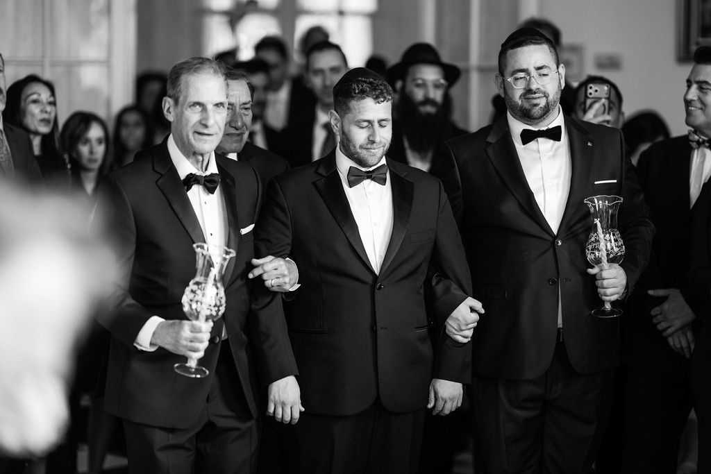 Black-and-white wedding celebration photo showing three men in black tuxedos holding ceremonial cups while smiling and walking past guests in the background.