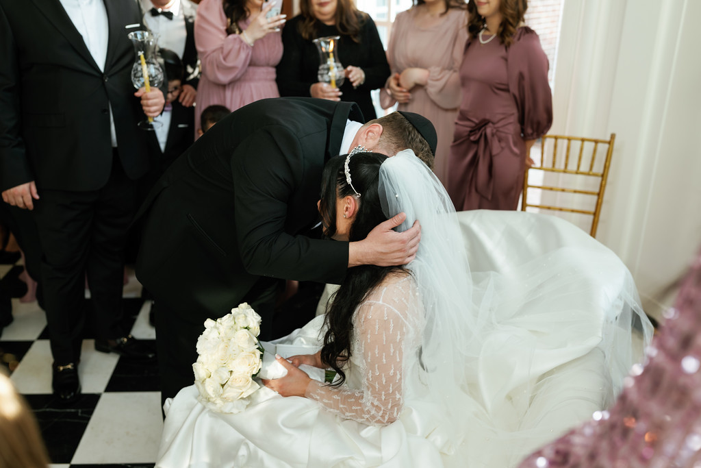 The groom bows close to the bride during the badeken, holding her gently as he lowers his head beside her veil while guests hold celebratory cups behind them.