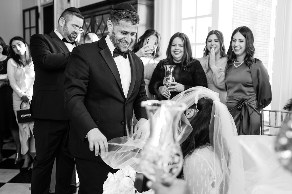 Black-and-white wedding reception moment: a bearded groom in a tuxedo smiles while guests in the background laugh and a bride and floral confetti fill the foreground.