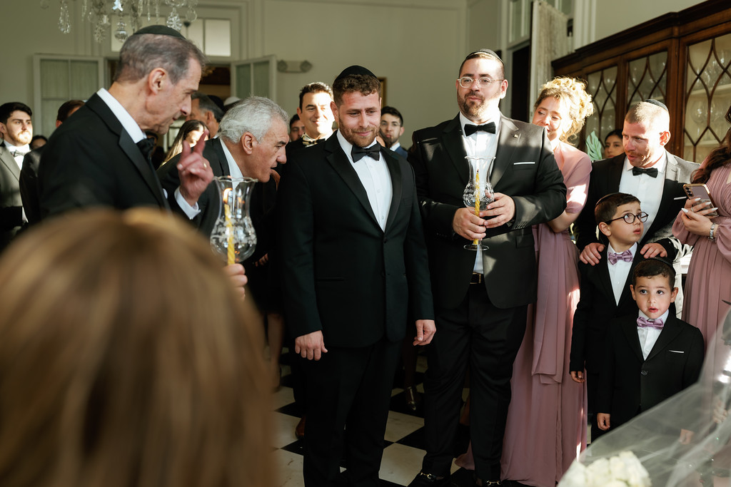 Groom and guests in black tuxedos standing together during kabbalas panim, with a groom holding a goblet and children nearby at a formal indoor event.