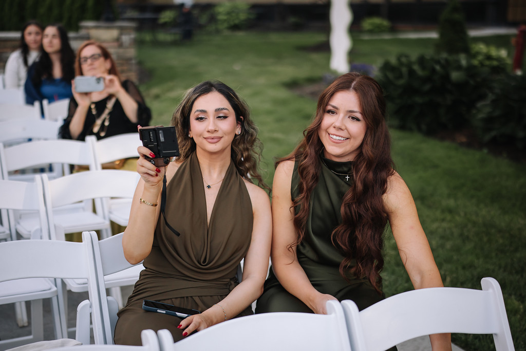 Two women seated on white chairs smiling at the camera during a wedding celebration, with other guests seated behind them on the lawn.