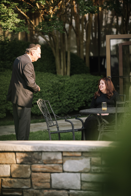 Outdoor candid of a laughing bride seated on a chair with the groom standing nearby during a quiet moment at a wedding.