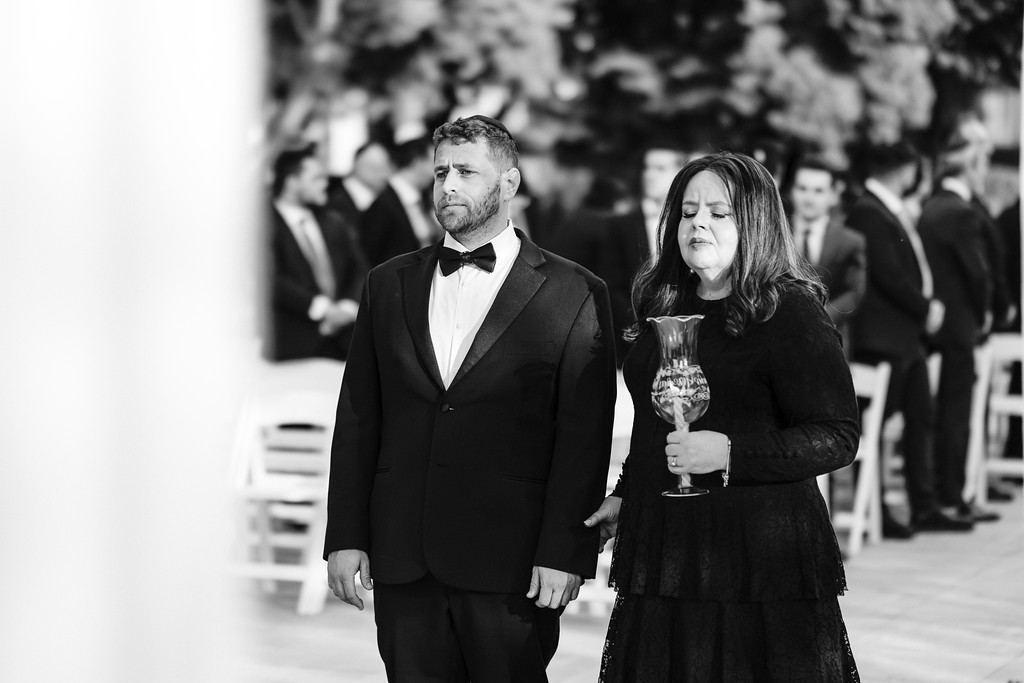 Black-and-white photo of a groom and his wife standing at their wedding venue, with the bride holding a glass and closing her eyes while the groom looks on.