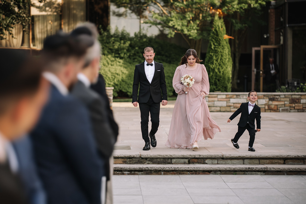 Outdoor wedding procession: the bride in a pale pink gown and the groom in a black suit walk down stone steps while two children run and smile toward them.