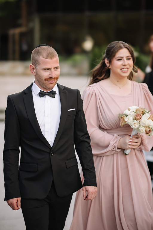 Groom in a black tuxedo and bow tie walking with the bride in a light pink gown holding a bouquet, outdoors during a wedding portrait moment.
