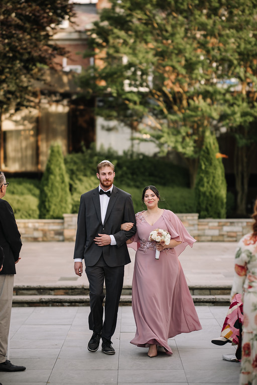 Framed in outdoor greenery, a bride in a mauve gown holds a bouquet while walking beside the groom in a black tuxedo during their wedding ceremony procession.