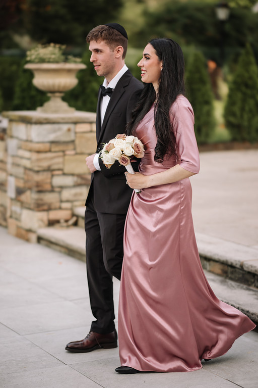 Outdoor couple portrait at a wedding: the bride in a mauve satin dress holding a bouquet and the groom in a black tuxedo standing beside her in a garden setting.