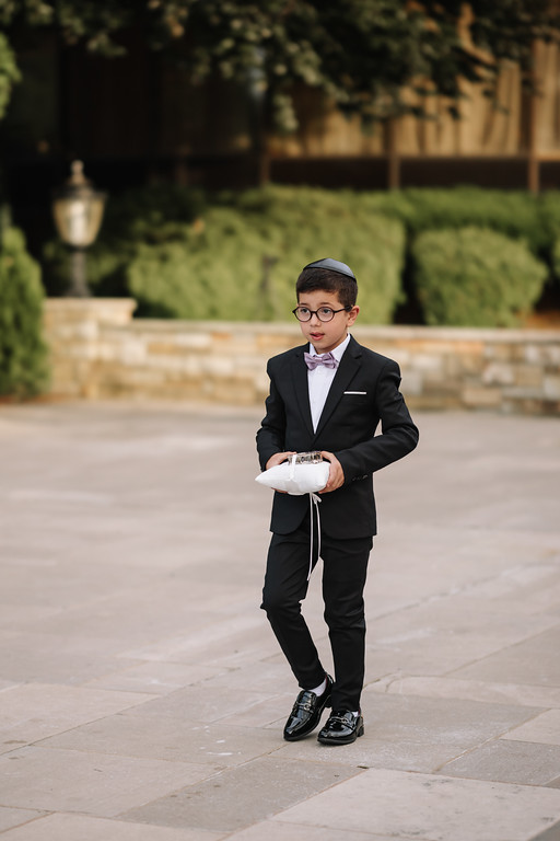 A young boy in a black suit with a bow tie and kippah stands outdoors on a stone walkway holding a small white item, looking directly at the camera.