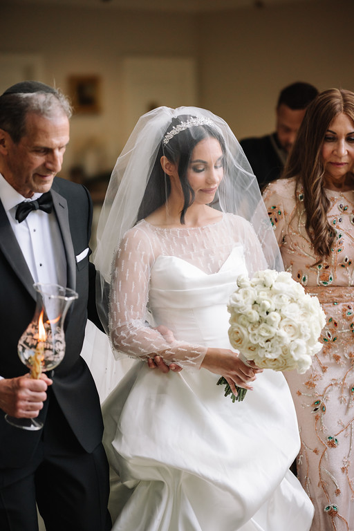 Frum bride in a white gown and lace veil with eyes closed as she holds a wedding bouquet, standing beside her father in a dark suit and bow tie.