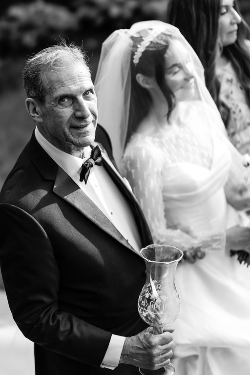 Black-and-white photo of the groom holding a ceremonial cup in the foreground with the bride in a bridal veil softly in the background.