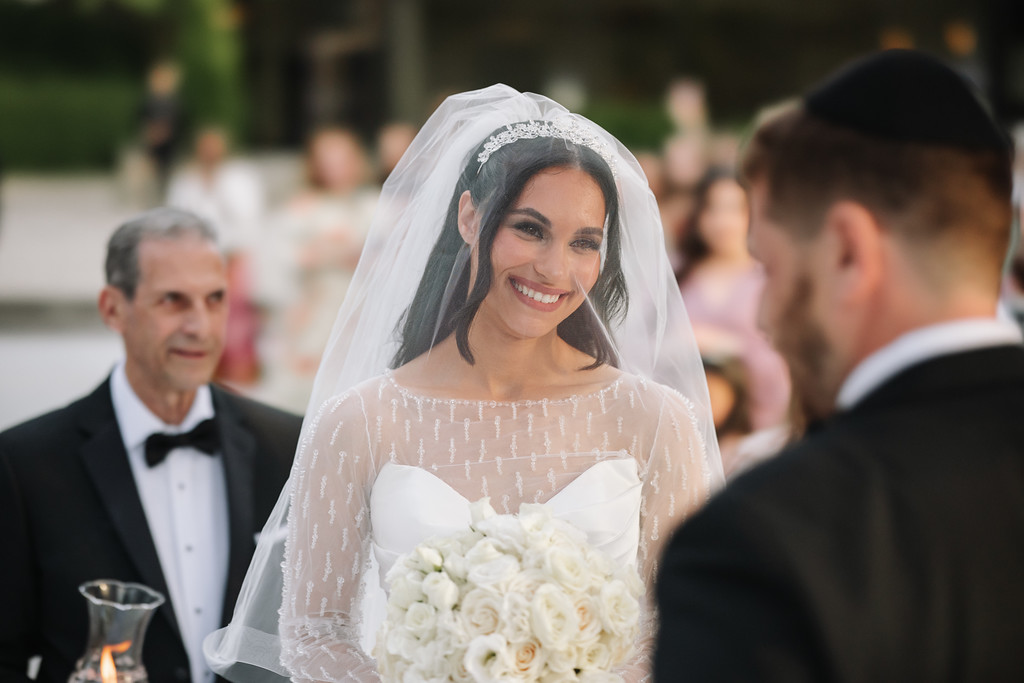 Frum bride smiling outdoors in a white veil while holding a bouquet, with guests in the background during the wedding ceremony.