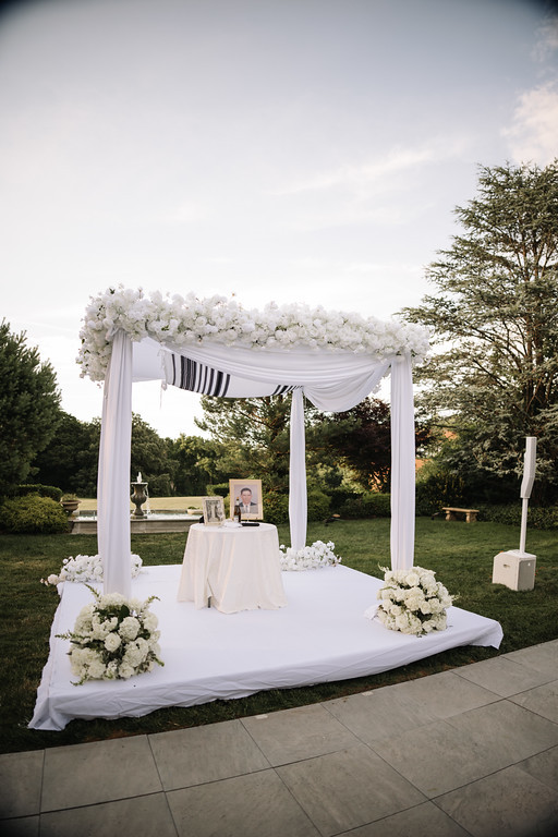 Outdoor chuppah setup on a garden lawn with white draped canopy, floral arrangements, and ceremony table in front of trees at dusk.