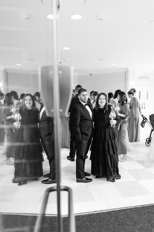 Black-and-white candid of a couple posing together at a reception as guests mingle behind them in a bright hall.