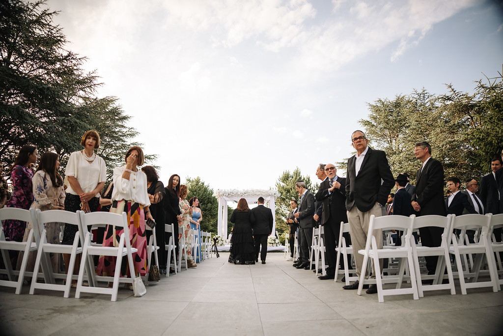 Guests and family line the walkway beside a white chuppah at an outdoor wedding, with the ceremony aisle stretching into the distance.